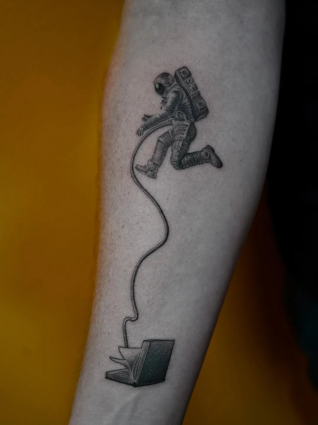 Astronaut floating from an open book, fine black and grey forearm tattoo